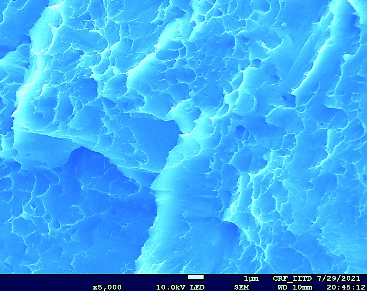 Sea of Fracture — false-colour SEM of a cleavage fracture surface in a Mg–Ca alloy, revealing river marks and terraced crack propagation. CRF IIT Delhi, ×5000.